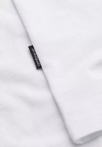 Hvid bomulds t-shirt med korte ærmer, prydet med et syet sort mærke på kanten. Glat tekstur, enkelt design, ingen mønstre eller grafik.
