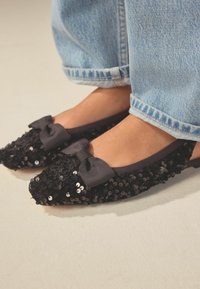 Ballerines noires en sequins avec un bout pointu, une bordure arrondie, et un détail en satin gris sur le devant, assorties à un jean en denim bleu clair.