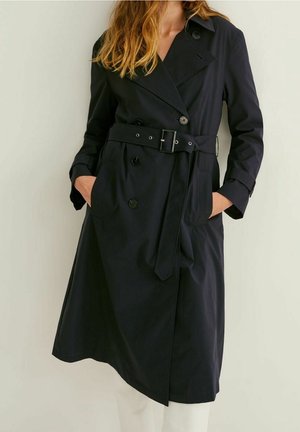 Un modèle porte un long trench-coat noir croisé avec une ceinture à la taille et les mains dans les poches.
