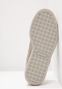 Suela de zapatilla beige con un agarre texturizado en forma de estrella y un borde gris liso. Logotipo exhibido en el centro de la suela.