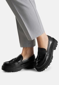 Černé kožené loafers se silnou grip podrážkou, s frendou a hladkým povrchem. Nosené s šedými kalhotami na míru.