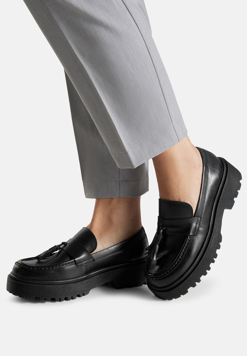 Černé kožené loafers se silnou grip podrážkou, s frendou a hladkým povrchem. Nosené s šedými kalhotami na míru.