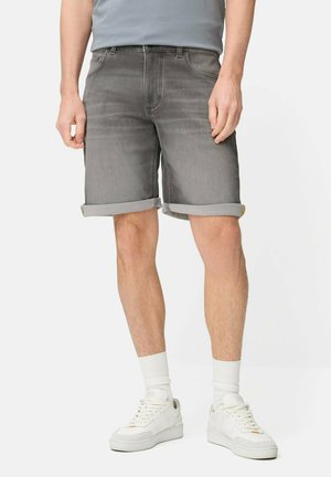 Man met grijze opgerolde spijkershorts, witte sportsokken, witte sneakers en een lichtgrijs ingestopt t-shirt, staand op witte achtergrond.