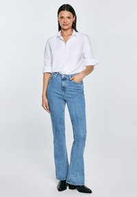 Žena stojící s rukou v kapse, oblečená v bílé košili na knoflíky, světle modrých džínách s vysokým pasem a černých kotníkových botách na jednobarevném pozadí.