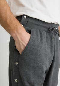 Szürke melegítőnadrág, zsinóros derékkal, oldalsó zsebekkel és patentos részletekkel a külső varráson. Puha, texturált anyagból készült.