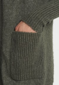 Maglione lavorato a maglia verde oliva con polsini a costine e una grande tasca testurizzata sulla parte anteriore, realizzato in morbido materiale di lana spazzolata.