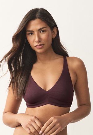 Žena s dugom tamnom kosom u bezrukom bordo braletu stoji s rukama lagano sklopljenim na jednostavnoj bijeloj pozadini.