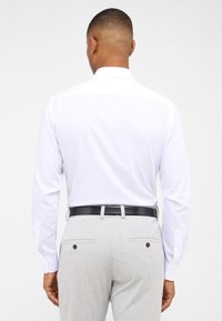 Witte blouse met lange mouwen en een standaard kraag, gecombineerd met lichtgrijze broek met twee achterzakken en een zwarte riem. Gladde stofstructuur.