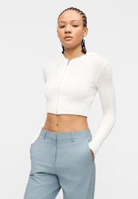 Νεαρό άτομο με πλεξούδες, φοράει ένα κομμένο λευκό φούτερ με φερμουάρ και ανοιχτό μπλε παντελόνι ραμμένο, με τα χέρια στις τσέπες, μπροστά σε απλό φόντο.