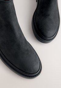 Paar schwarze Wildleder-Stiefeletten mit abgerundeten Zehen und dicken Gummisohlen auf hellem Untergrund.