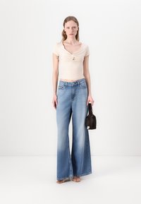 Top fitted beige chiaro con dettaglio a torsione, abbinato a jeans in denim blu a gamba larga e una piccola borsa nera. Texture morbide, stile casual.