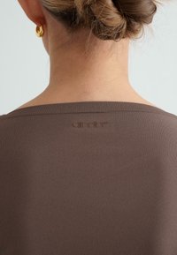 Smeđa majica s rebrastim izrezom i suptilnim logotipom brenda izvezenim na leđima. Glatka tekstura, minimalistički dizajn.