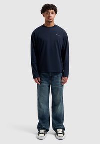 Marineblaues Langarm-T-Shirt mit Rundhalsausschnitt, kombiniert mit hellblauen Jeans mit geradem Bein und schwarzen Sneakern mit weißen Sohlen.