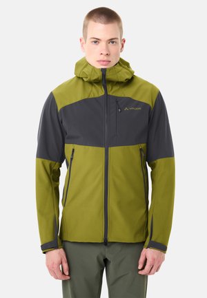 Uomo che indossa una giacca outdoor verde e grigio scuro con cappuccio, cerniera frontale e tasche con zip, in piedi davanti a uno sfondo semplice.