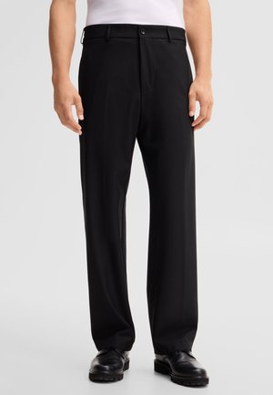Uomo che indossa pantaloni neri a gamba dritta, scarpe eleganti nere e una camicia bianca infilata, in piedi su uno sfondo chiaro e semplice.