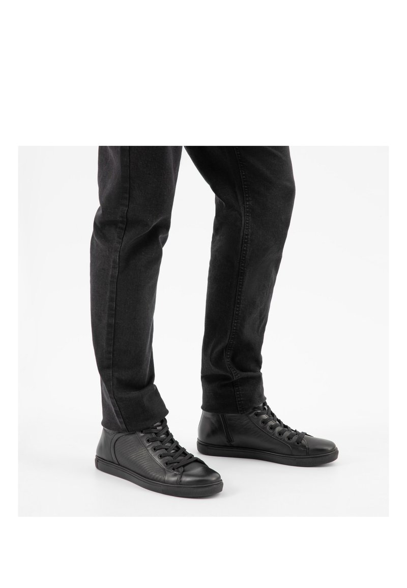 Osoba stojící na bílém pozadí, obutá do černých vysokých kožených tenisek a černých džínů.
