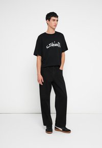 Juoda trumpomis rankovėmis marškinėliai su balta grafine užrašu, derinami su juodomis platėjusiomis kelnėmis ir juodais sportbačiais su rudos spalvos padais.