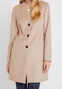 Beige uld blandingsfrakke med spidse kraver, fem sorte knapper og sidelommer. Glat tekstur med skræddersyet snit og knælængde design.