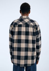 Camisa de franela a cuadros en negro y beige con un corte relajado, mangas largas y puños abotonados, con un dobladillo curvado y detalle en la yugo trasero.