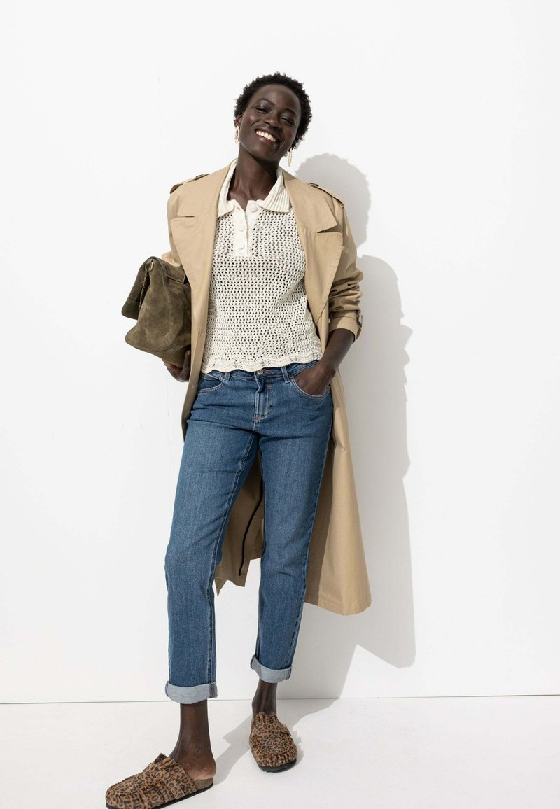 Manteau trench beige, haut en tricot blanc, jeans bleus retroussés, et chaussures à enfiler en imprimé léopard marron. Accessoire : sac vert olive.