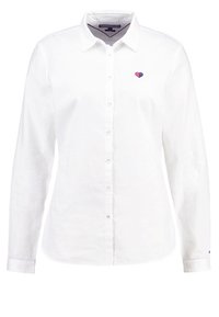 Chemise blanche à manches longues avec col et petit emblème en forme de cœur rose et bleu sur la poitrine gauche.