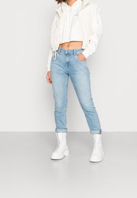 Witte cropped hoodie gecombineerd met een beige gewatteerd vest, lichtblauwe spijkerbroek en chunky witte enkellaarsjes. Heeft een relaxte pasvorm en opgerolde manchetten.