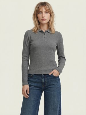 Junge Frau mit schulterlangen blonden Haaren, die ein eng anliegendes graues Langarm-Poloshirt und blaue Jeans trägt, steht mit einer Hand in der Tasche.
