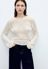Pulover tricotat în crem, cu un model de țesătură deschisă și mâneci lungi, asortat cu blugi din denim de un albastru închis. Design simplu și relaxat.