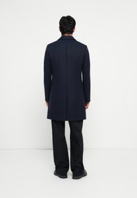 Palton lung din lână bleumarin cu un design ajustat, având o siluetă dreaptă, rever în formă de V și manșete cu butoane, asortat cu pantaloni negri.