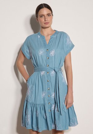 Vrouw in een lichtblauwe jurk met knopen aan de voorkant, witte bloemenborduursels en een strikceintuur, staand met één hand in de zak.