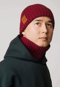 Bordowa dzianinowa czapka z teksturowanym, prążkowanym wzorem oraz skórzaną naszywką z logo, noszona z ciemnozielonym kapturem.