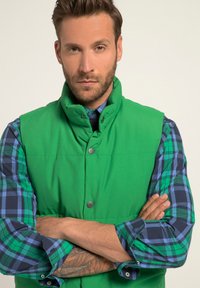 Zielona kamizelka ocieplana z wysokim kołnierzem i zatrzaskami, noszona na niebieskiej i zielonej koszuli w kratkę z guziczkami przy mankietach.