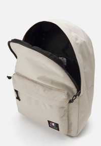Beige fabric backpack with a rounded top, front zip pocket, and inner black lining. Features logo patch and drawstring closure.