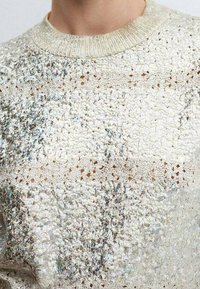 Suéter de tono plateado de punto con un patrón texturizado, que presenta un cuello acanalado y acentos brillantes en toda la tela.