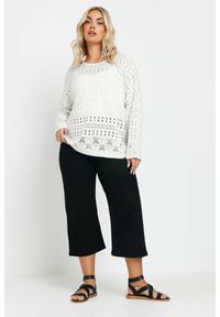 Vit stickad tröja med perforerade mönster, tillsammans med svarta cropped byxor och svarta sandaletter med remmar. Släta texturer och avslappnad passform.