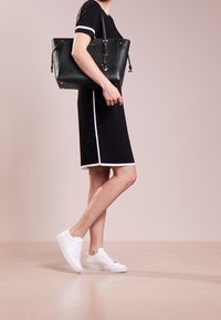 Mujer vistiendo un vestido negro con ribetes blancos y zapatillas blancas, llevando un gran bolso negro sobre su hombro frente a un fondo neutro.