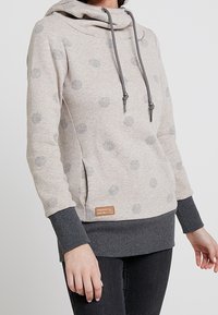 Beige hoodie med gråa prickar, mörkgrå muddar och fåll, framficka och huva med snören, bärs med svarta byxor.