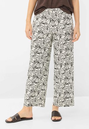 Persona che indossa pantaloni ampi con stampa di foglie in bianco e nero e sandali neri aperti, in piedi contro uno sfondo bianco.