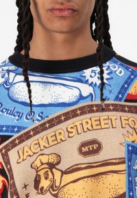 Graphic sweater featuring a multicolored design with text. Material appears knitted, with blue, red, and beige sections. Accents include star patterns.