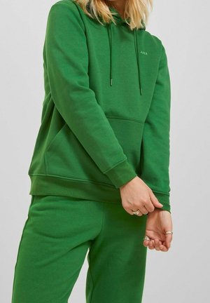 Osoba ubrana w zieloną bluzę z kapturem i dopasowane spodnie dresowe, poprawiająca rękaw z dłońmi ozdobionymi srebrnymi pierścieniami na jednolitym tle.