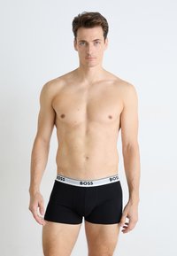 Schwarze Boxershorts mit einem weißen Bund, der das "BOSS"-Logo zeigt. Das Material wirkt glatt und elastisch, im taillierten Design. Das Modell zeigt einen neutralen Gesichtsausdruck.