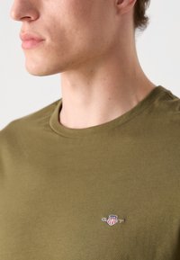 T-shirt en coton vert olive avec un col rond et un petit logo brodé sur la poitrine. Texture lisse, design décontracté.