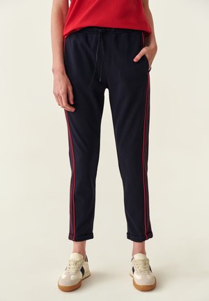 Pantaloni jogger blu navy con strisce rosse ai lati, vita con coulisse e orli arrotolati. Abbinati a sneakers bianche con suola beige e accenti blu navy.