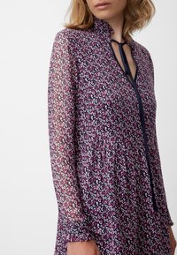 Blommönstrad klänning i lila, med långa genomskinliga ärmar, hög krage och en knytning vid halsen samt en samlad midja.