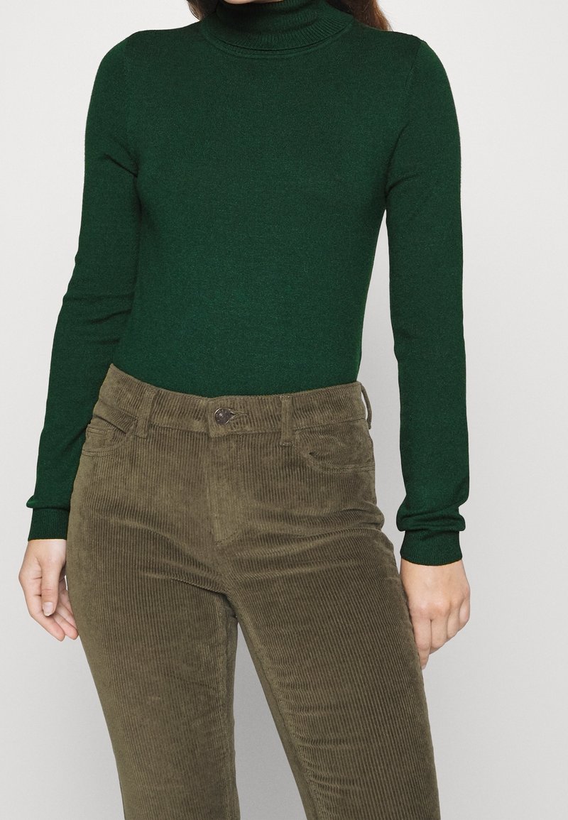 Personne portant un pull à col roulé vert foncé et un pantalon en velours côtelé vert olive taille haute, debout devant un fond uni.