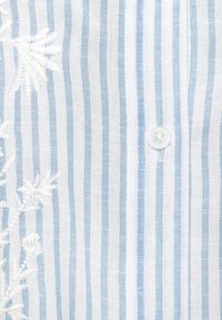 Primer plano de tela con rayas verticales blancas y azul claro, bordado floral blanco y un pequeño botón redondo blanco de cuatro orificios.