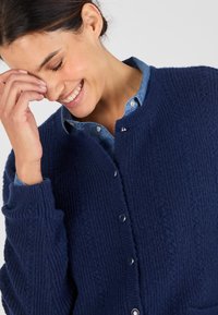 Cardigan lavorato a maglia blu navy con una texture a coste, caratterizzato da bottoni argentati e un collo rotondo. Indossato sopra una camicia azzurro chiaro.