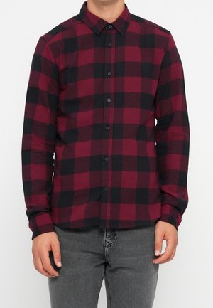 Chemise en flanelle rouge et noire à motif à carreaux, manches longues, col boutonné et boutons noirs, portée avec un jean gris. Texture douce et coupe régulière.