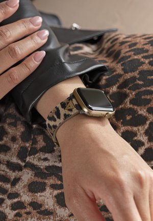 Približen posnetek zapestja z žepno uro z leopardovim tiskom pasu, ki počiva na tkanini z enakim leopardovim tiskom, z roko, ki se dotika črnega usnjenega rokava.