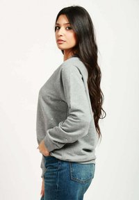 Felpa grigia con vestibilità ampia, decorata con piccoli e delicati ornamenti. Maniche lunghe, tessuto leggero, abbinata a jeans blu.